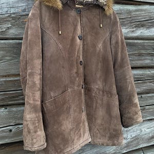 May include: A brown suede coat with a fur-lined hood and button closure. The coat has two front pockets and a tailored design. The hood has a fur trim. The coat is hanging on a wooden hanger.
