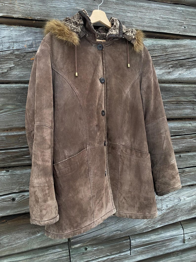 May include: A brown suede coat with a fur-lined hood and button closure. The coat has two front pockets and a tailored design. The hood has a fur trim. The coat is hanging on a wooden hanger.