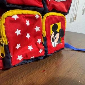 May include: Red and blue backpack with yellow zippers and white star pattern. Features a Mickey Mouse graphic on a front pocket. The backpack has a blue strap.