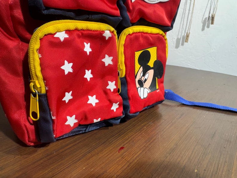 May include: Red and blue backpack with yellow zippers and white star pattern. Features a Mickey Mouse graphic on a front pocket. The backpack has a blue strap.