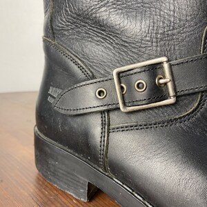 May include: Black leather ankle boots with a buckle strap and a silver buckle. The boots have a stitched seam around the ankle and a thick sole.