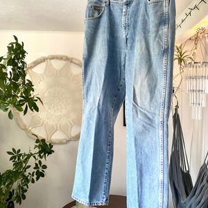 May include: Light blue denim jeans hanging on a wooden hanger. The jeans have a classic five-pocket design, a button and zipper closure, and a straight leg. The jeans are made of a faded denim material.