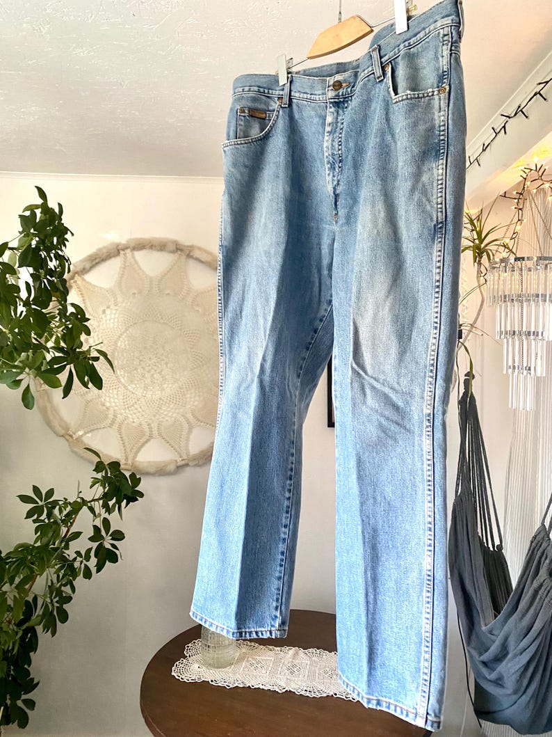 May include: Light blue denim jeans hanging on a wooden hanger. The jeans have a classic five-pocket design, a button and zipper closure, and a straight leg. The jeans are made of a faded denim material.