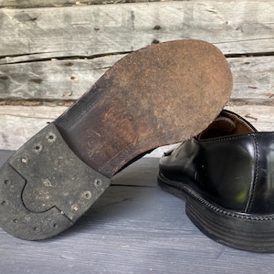 May include: A pair of black leather dress shoes. One shoe is flipped over, revealing the worn sole with metal studs. The other shoe is upright, showcasing the shiny, polished leather and the sole's textured design.