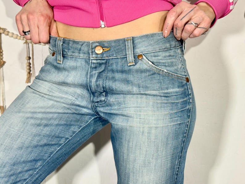 May include: Light blue denim jeans with a classic five-pocket design. The jeans have a button and zipper closure, and copper-colored rivets. The denim has a faded wash, giving it a worn-in look. The jeans are a mid-rise style.