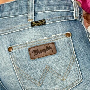 May include: Close-up of light blue denim jeans with a distressed pocket. The Wrangler brand is visible on a brown leather patch on the back pocket. The jeans have a classic five-pocket design and copper-colored rivets.
