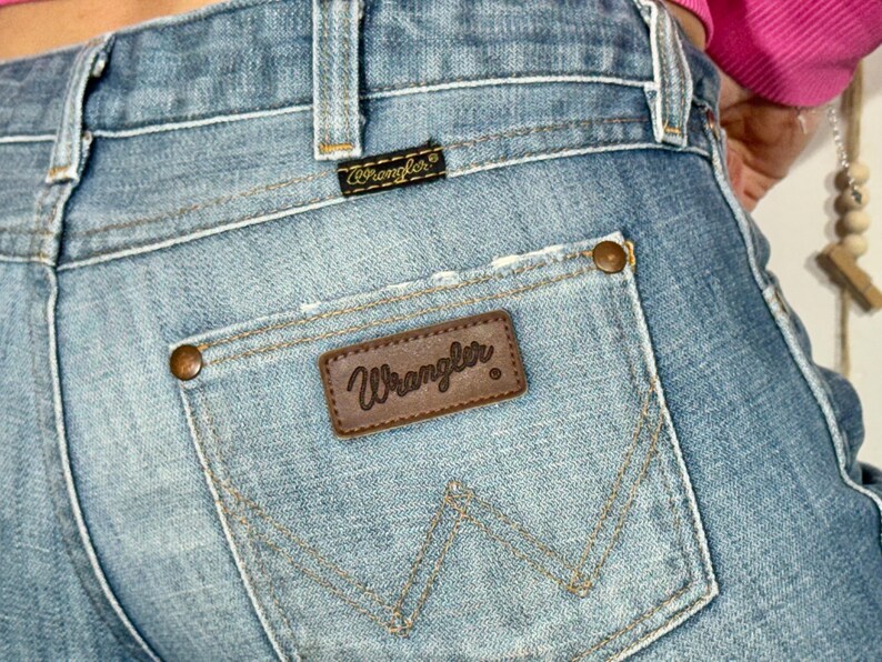 May include: Close-up of light blue denim jeans with a distressed pocket. The Wrangler brand is visible on a brown leather patch on the back pocket. The jeans have a classic five-pocket design and copper-colored rivets.