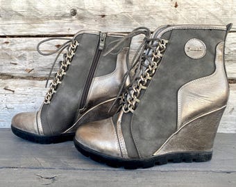 Puede incluir: Un par de botines de cuña grises y plateados con un detalle de cadena en los cordones. Los botines tienen una cremallera en el interior del tobillo y un nombre de marca en el lateral.