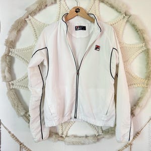 May include: White Fila track jacket with black trim and a red and white logo on the chest. The jacket has a full zipper and is hanging on a wooden hanger. A vintage tennis racket is visible on the left.