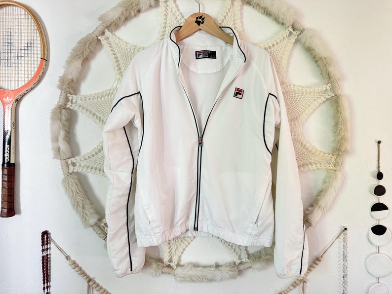 May include: White Fila track jacket with black trim and a red and white logo on the chest. The jacket has a full zipper and is hanging on a wooden hanger. A vintage tennis racket is visible on the left.