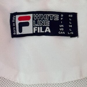 May include: Close-up of a white garment with a black label. The label features the FILA logo in red, white, and black, along with the text "WHITE LINE FILA." Additional text includes "Born in Italy" and size indicators.