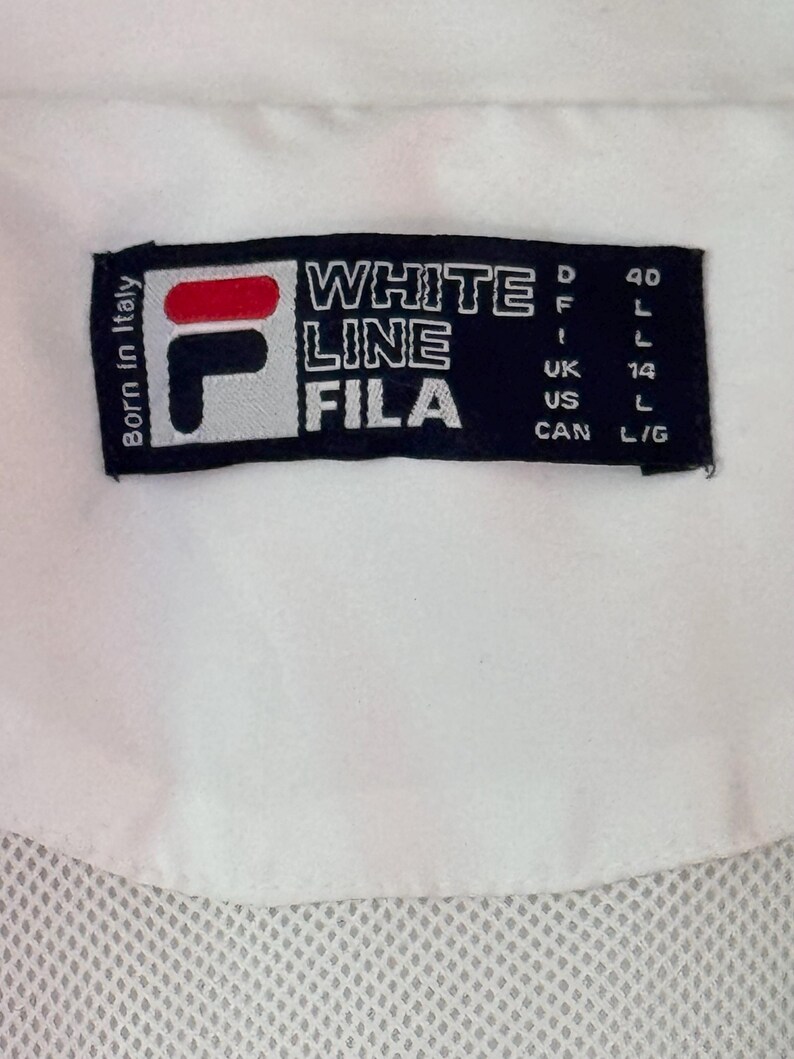 May include: Close-up of a white garment with a black label. The label features the FILA logo in red, white, and black, along with the text "WHITE LINE FILA." Additional text includes "Born in Italy" and size indicators.