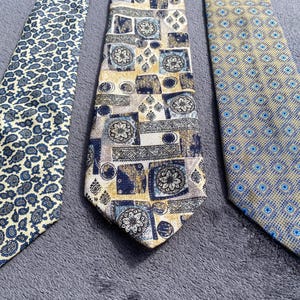 Puede incluir: Tres corbatas estampadas. La primera corbata tiene un fondo crema con un diseño de cachemira azul. La segunda corbata tiene un fondo beige con un patrón geométrico en azul marino, crema y gris. La tercera corbata tiene un fondo azul claro con un patrón de diamantes azul y crema repetido.