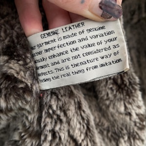 May include: Close-up of a tag that reads "GENUINE LEATHER" in black text. The tag is white and attached to a brown and gray faux fur material. The text on the tag describes the garment's genuine leather composition and natural imperfections.