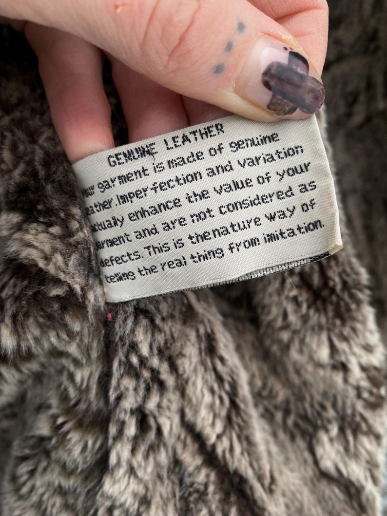 May include: Close-up of a tag that reads "GENUINE LEATHER" in black text. The tag is white and attached to a brown and gray faux fur material. The text on the tag describes the garment's genuine leather composition and natural imperfections.