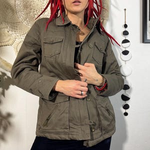 May include: An olive green jacket featuring multiple pockets, a button-up front, and a collared neckline. The jacket has a military-inspired design and a slightly worn appearance. The sleeves are long, and the jacket appears to be made of a durable fabric.