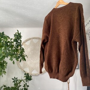 May include: A brown sweater hangs on a wooden hanger. The sweater has a crew neck and long sleeves. The material appears to be a soft knit. The background includes a plant, a decorative wall hanging, and a light fixture.