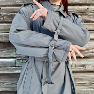 May include: A gray trench coat with a belted waist and adjustable sleeve straps. The coat features a classic collar and a double-breasted design. The fabric appears to be a lightweight material, suitable for transitional weather. The background is a wooden wall.