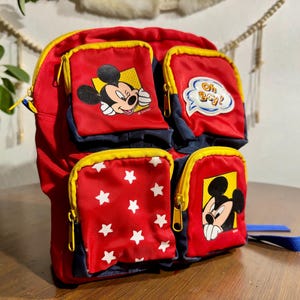 May include: Red and navy blue backpack featuring Mickey Mouse graphics and star patterns. The backpack has multiple zippered compartments with yellow trim and a blue strap. One pocket has the text "Oh Boy!"