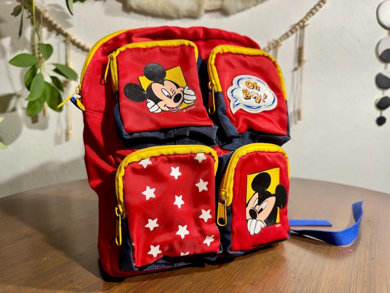 May include: Red and navy blue backpack featuring Mickey Mouse graphics and star patterns. The backpack has multiple zippered compartments with yellow trim and a blue strap. One pocket has the text "Oh Boy!"