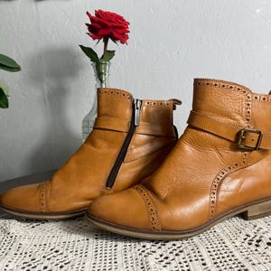 K&ouml;nnte beinhalten: Ein Paar hellbraune Leder-Ankle Boots mit dekorativen N&auml;hten und Schnallen. Ein Stiefel hat einen seitlichen Rei&szlig;verschluss. Die Stiefel stehen auf einem Tisch mit einem Spitzentischl&auml;ufer, mit einer roten Rose in einer Vase im Hintergrund.