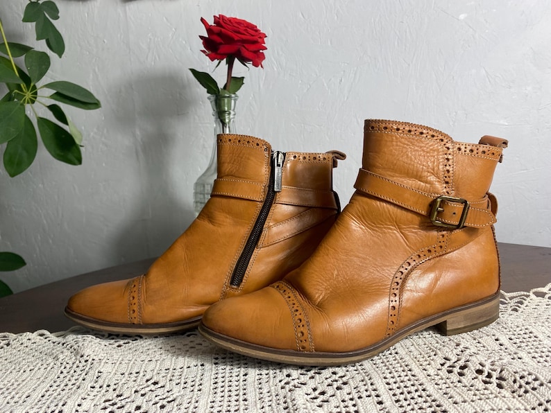 K&ouml;nnte beinhalten: Ein Paar hellbraune Leder-Ankle Boots mit dekorativen N&auml;hten und Schnallen. Ein Stiefel hat einen seitlichen Rei&szlig;verschluss. Die Stiefel stehen auf einem Tisch mit einem Spitzentischl&auml;ufer, mit einer roten Rose in einer Vase im Hintergrund.