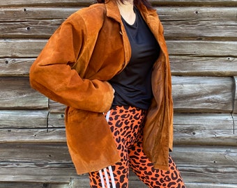 May include: A brown suede jacket with a long, loose fit. The jacket has a button-up front and a collar. The person wearing the jacket is also wearing leopard print leggings with white stripes.