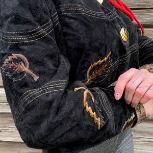 May include: A black suede jacket with gold stitching and brown embroidered leaves. The jacket has a button closure and a stand-up collar.