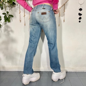 May include: Light blue denim jeans with a Wrangler label on the back pocket. The jeans have a classic five-pocket design and a straight leg fit. The jeans are worn with white platform sneakers.