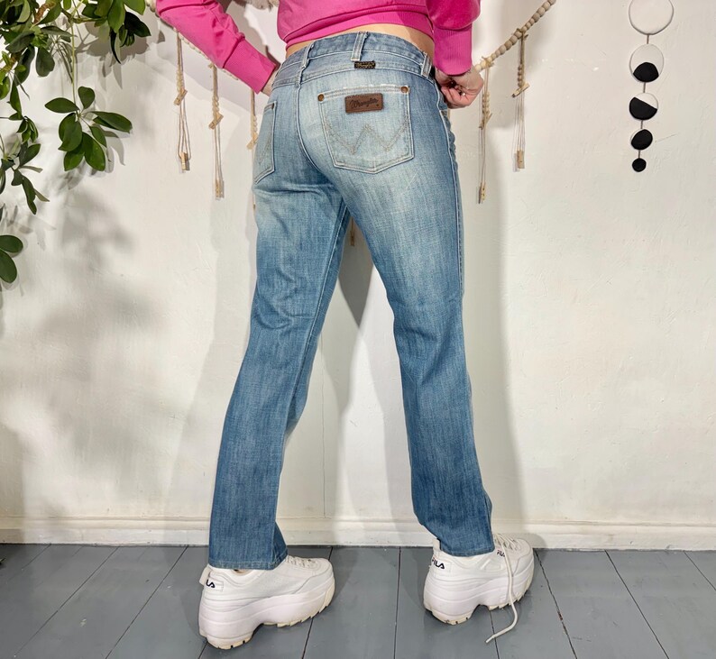 May include: Light blue denim jeans with a Wrangler label on the back pocket. The jeans have a classic five-pocket design and a straight leg fit. The jeans are worn with white platform sneakers.