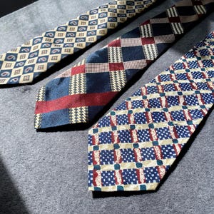 Könnte beinhalten: Drei gemusterte Krawatten nebeneinander. Die Krawatten haben verschiedene Designs, darunter Paisley-, Karo- und geometrische Muster. Die Farben sind Marineblau, Rot, Beige und Weiß. Die Krawatten liegen auf einer grauen Oberfläche.