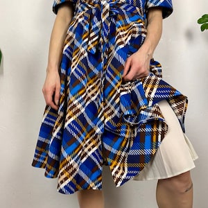 May include: A blue, brown, and white plaid wrap dress with a white slip underneath. The dress has a cinched waist and a flowing skirt.