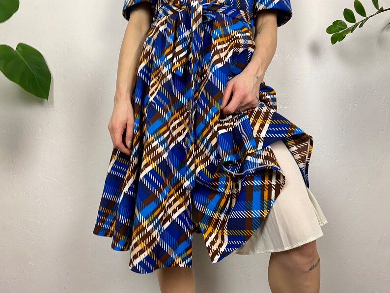 May include: A blue, brown, and white plaid wrap dress with a white slip underneath. The dress has a cinched waist and a flowing skirt.