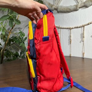 May include: Red backpack with blue and yellow accents. The backpack has a main compartment and two side pockets with yellow zippers. Blue shoulder straps and a top handle complete the design. The backpack is made of a durable material.