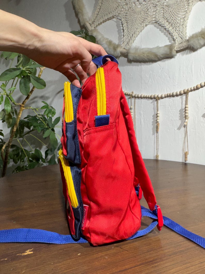May include: Red backpack with blue and yellow accents. The backpack has a main compartment and two side pockets with yellow zippers. Blue shoulder straps and a top handle complete the design. The backpack is made of a durable material.