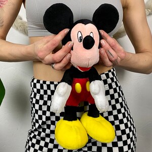 May include: A plush Mickey Mouse toy with black ears, a red shirt, yellow shoes, and a black nose. The toy is being held by a person wearing a gray tank top and black and white checkered pants.