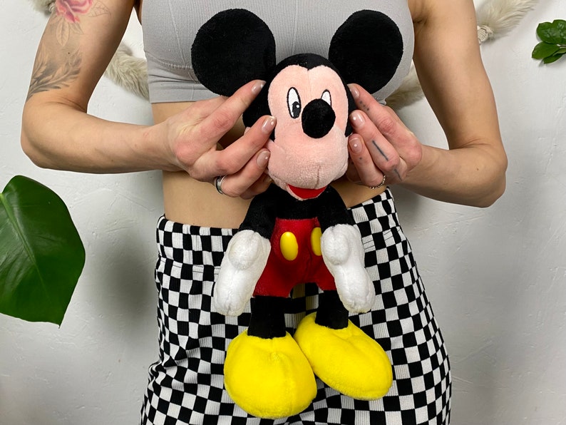 May include: A plush Mickey Mouse toy with black ears, a red shirt, yellow shoes, and a black nose. The toy is being held by a person wearing a gray tank top and black and white checkered pants.