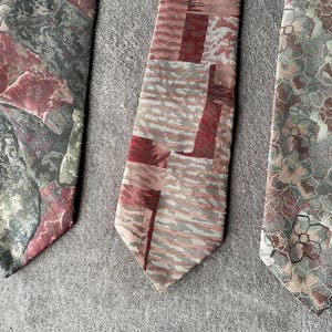 Könnte beinhalten: Drei gemusterte Krawatten in Grau-, Burgunder- und Grüntönen werden gezeigt. Die Krawatten haben einzigartige Designs, darunter abstrakte Formen und florale Muster. Die Krawatten sind auf einer grauen Oberfläche angeordnet.