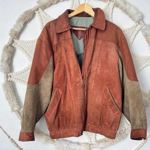 May include: A vintage leather jacket in shades of brown and tan, hanging on a wooden hanger. The jacket has a collared neckline, a zip closure, and two front pockets. The sleeves are a lighter tan colour.