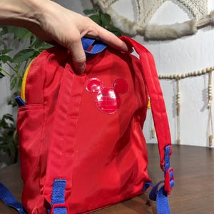 May include: Red backpack with a Mickey Mouse emblem. The backpack has blue straps and accents, a yellow zipper, and a small side pocket. The backpack is made of a shiny, durable material.