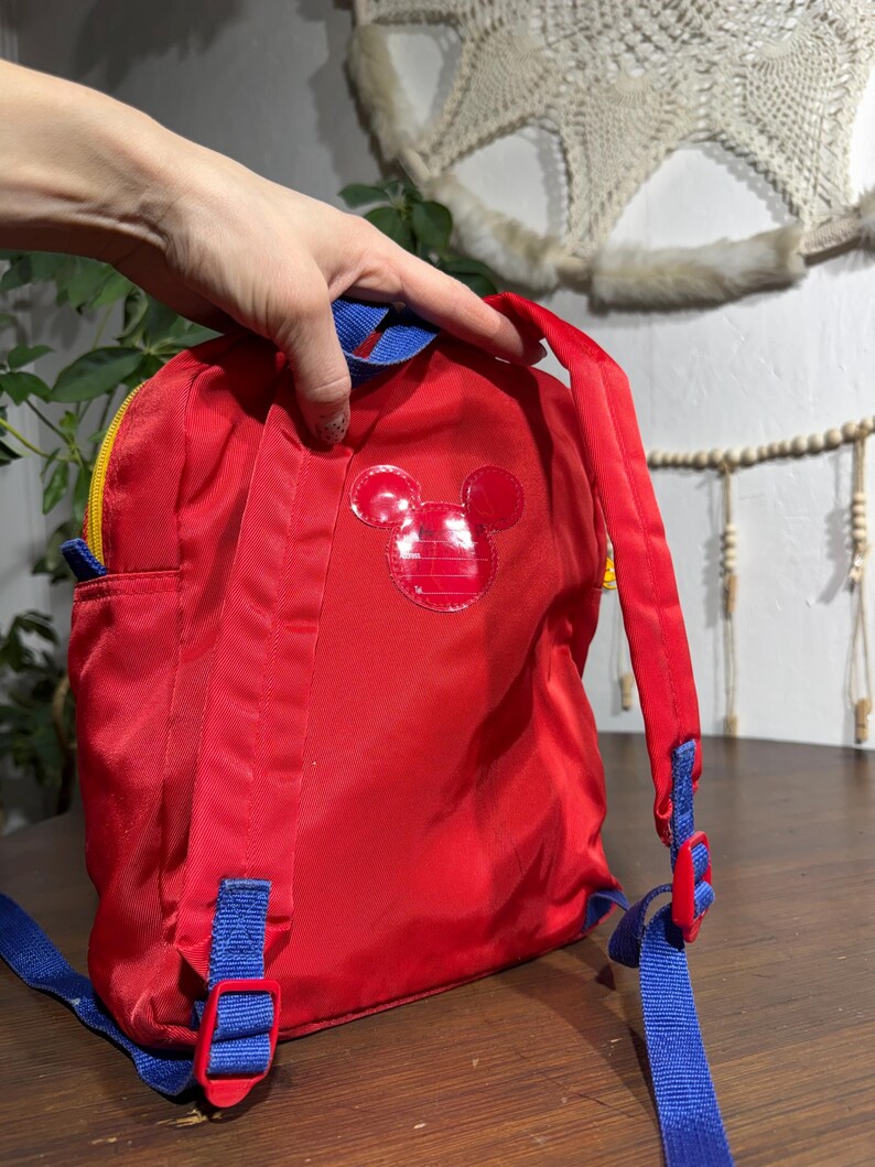 May include: Red backpack with a Mickey Mouse emblem. The backpack has blue straps and accents, a yellow zipper, and a small side pocket. The backpack is made of a shiny, durable material.