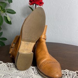 K&ouml;nnte beinhalten: Ein Paar hellbraune Leder-Ankle-Boots mit Schnallen. Die Stiefel haben eine runde Spitze und einen niedrigen Absatz. Die Sohlen sind hellbraun. Im Hintergrund ist eine rote Rose zu sehen.