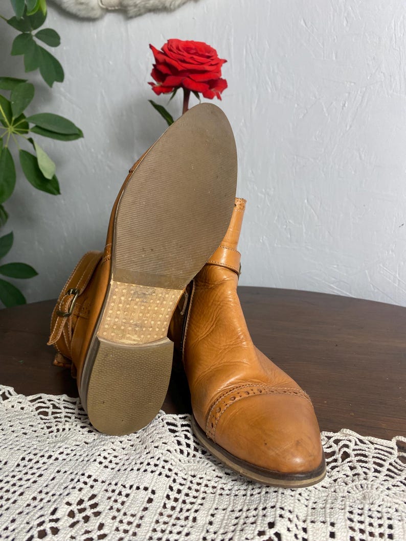 K&ouml;nnte beinhalten: Ein Paar hellbraune Leder-Ankle-Boots mit Schnallen. Die Stiefel haben eine runde Spitze und einen niedrigen Absatz. Die Sohlen sind hellbraun. Im Hintergrund ist eine rote Rose zu sehen.