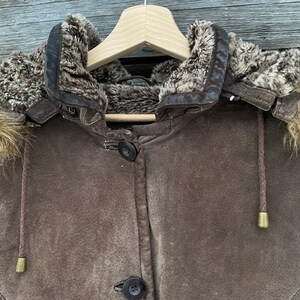 May include: A brown suede jacket with a fur-lined hood, hanging on a wooden hanger. The jacket features a button-up front and drawstrings with gold-colored tips. The hood has a fur trim and a leather collar.