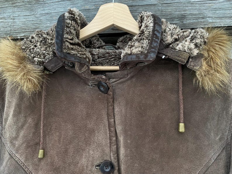 May include: A brown suede jacket with a fur-lined hood, hanging on a wooden hanger. The jacket features a button-up front and drawstrings with gold-colored tips. The hood has a fur trim and a leather collar.