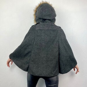 May include: A dark gray cape with a hood trimmed with faux fur. The hooded cape has a button closure and a wide, flowing design. The back view shows the cape's shape and details, suitable for cold weather.