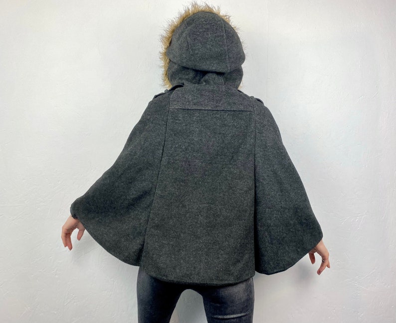 May include: A dark gray cape with a hood trimmed with faux fur. The hooded cape has a button closure and a wide, flowing design. The back view shows the cape's shape and details, suitable for cold weather.