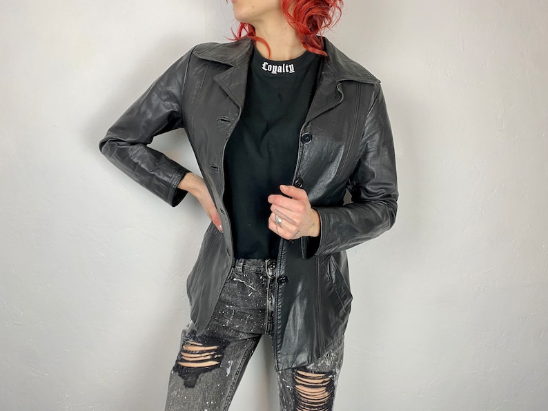 May include: A black leather jacket with a button closure and a collar. The jacket has a slim fit and is worn over a black t-shirt with the word "Loyalty" printed on it. The person is wearing distressed jeans.