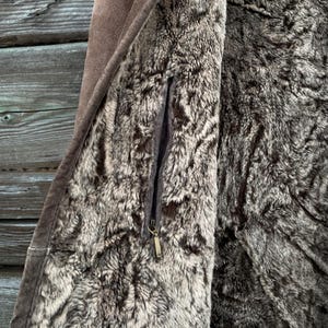 May include: Close-up of a brown suede coat with a faux fur lining. The coat features a zipper closure and a small pocket. The interior lining is a mix of gray and brown faux fur.