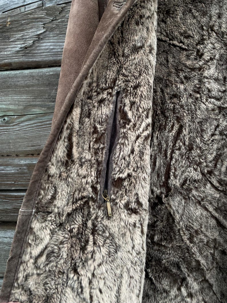 May include: Close-up of a brown suede coat with a faux fur lining. The coat features a zipper closure and a small pocket. The interior lining is a mix of gray and brown faux fur.